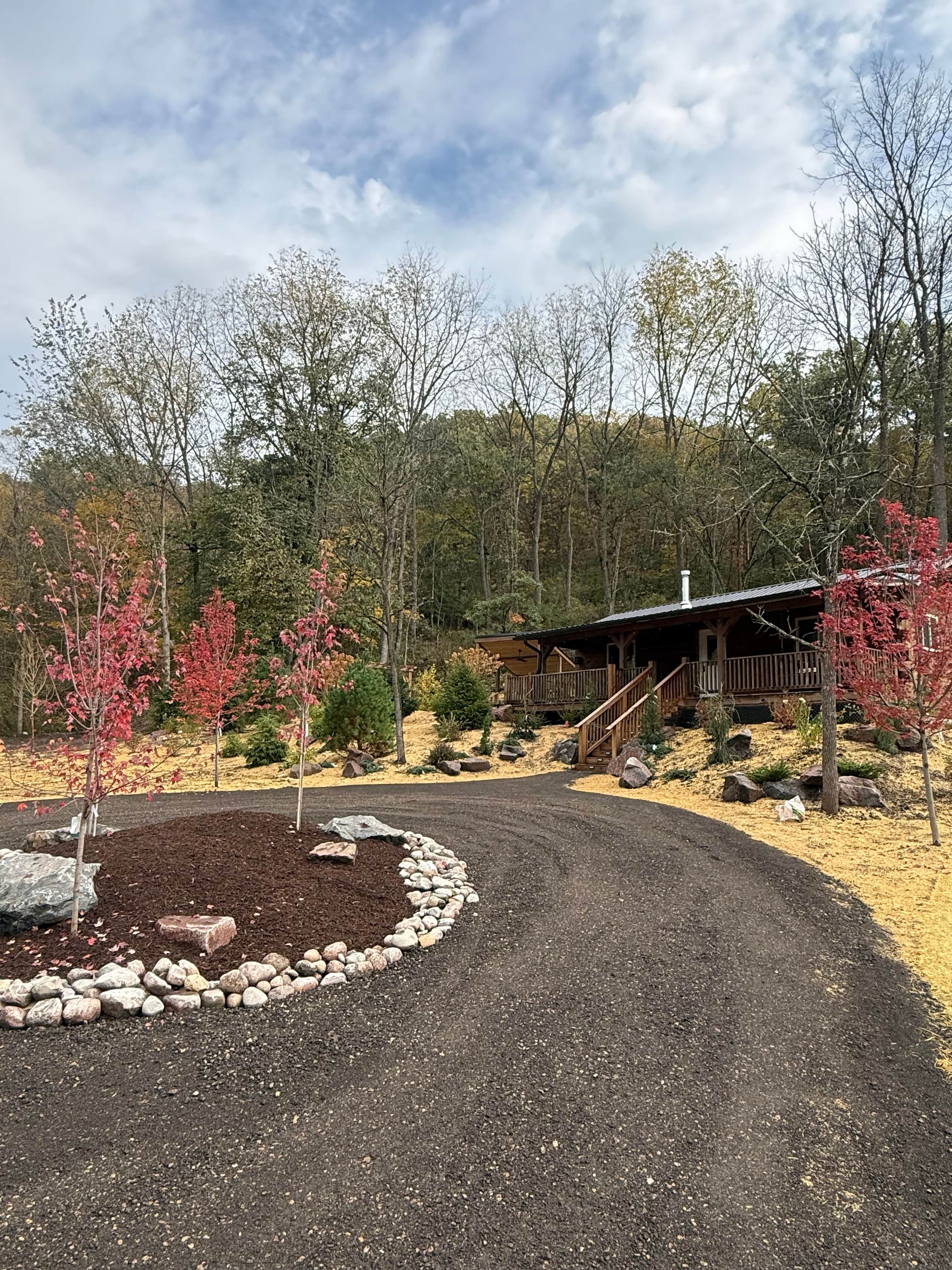 A gravel driveway leading to a wooden cabin with a covered porch, surrounded by trees and a landscaped area with rocks and shrubs.