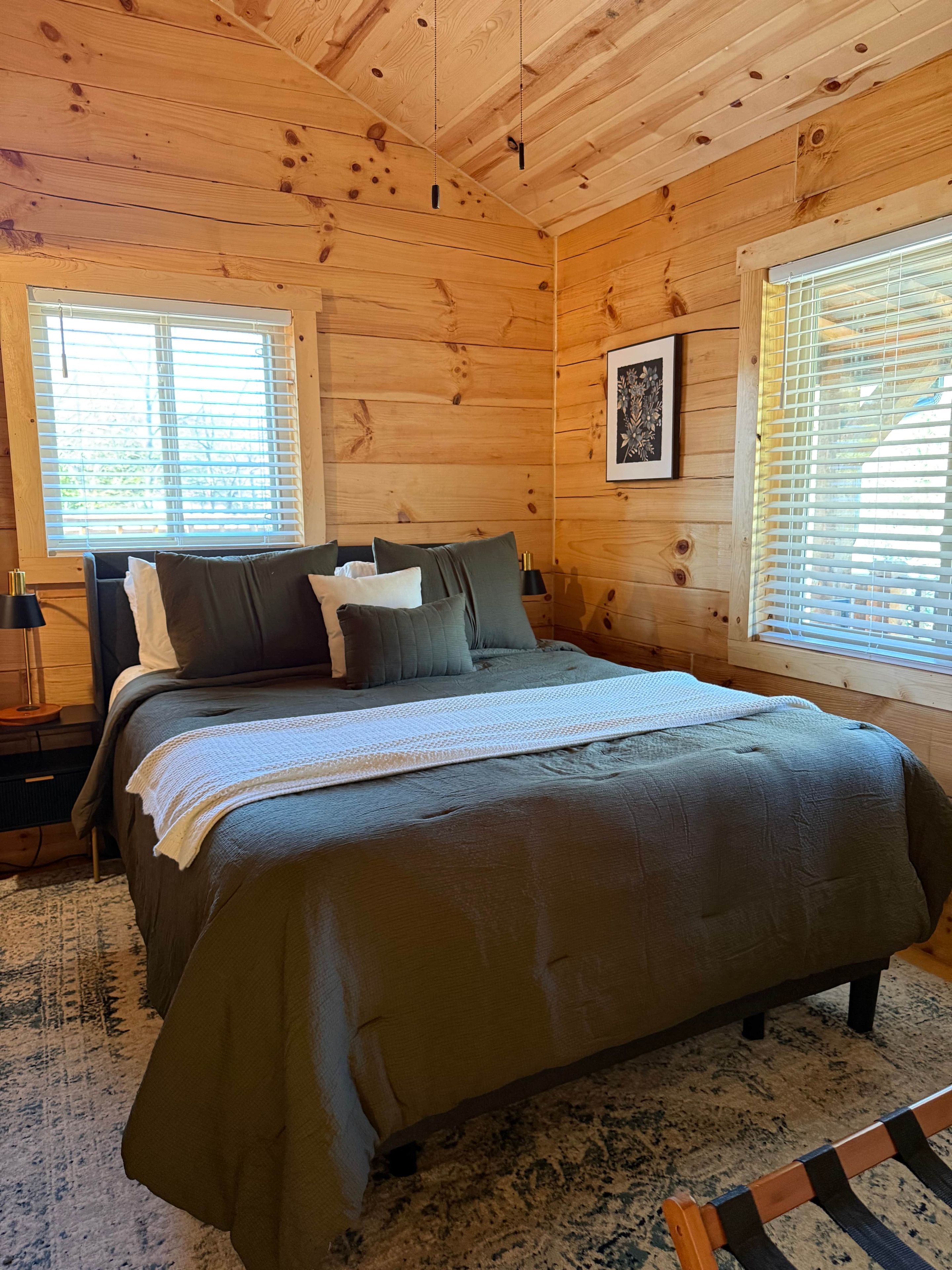 A wooden cabin bedroom with a bed, a nightstand, a framed picture, and a window with blinds.