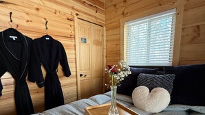 Une pièce de cabane en bois comprenant un lit, une petite table en bois avec un vase et une fenêtre avec des stores.