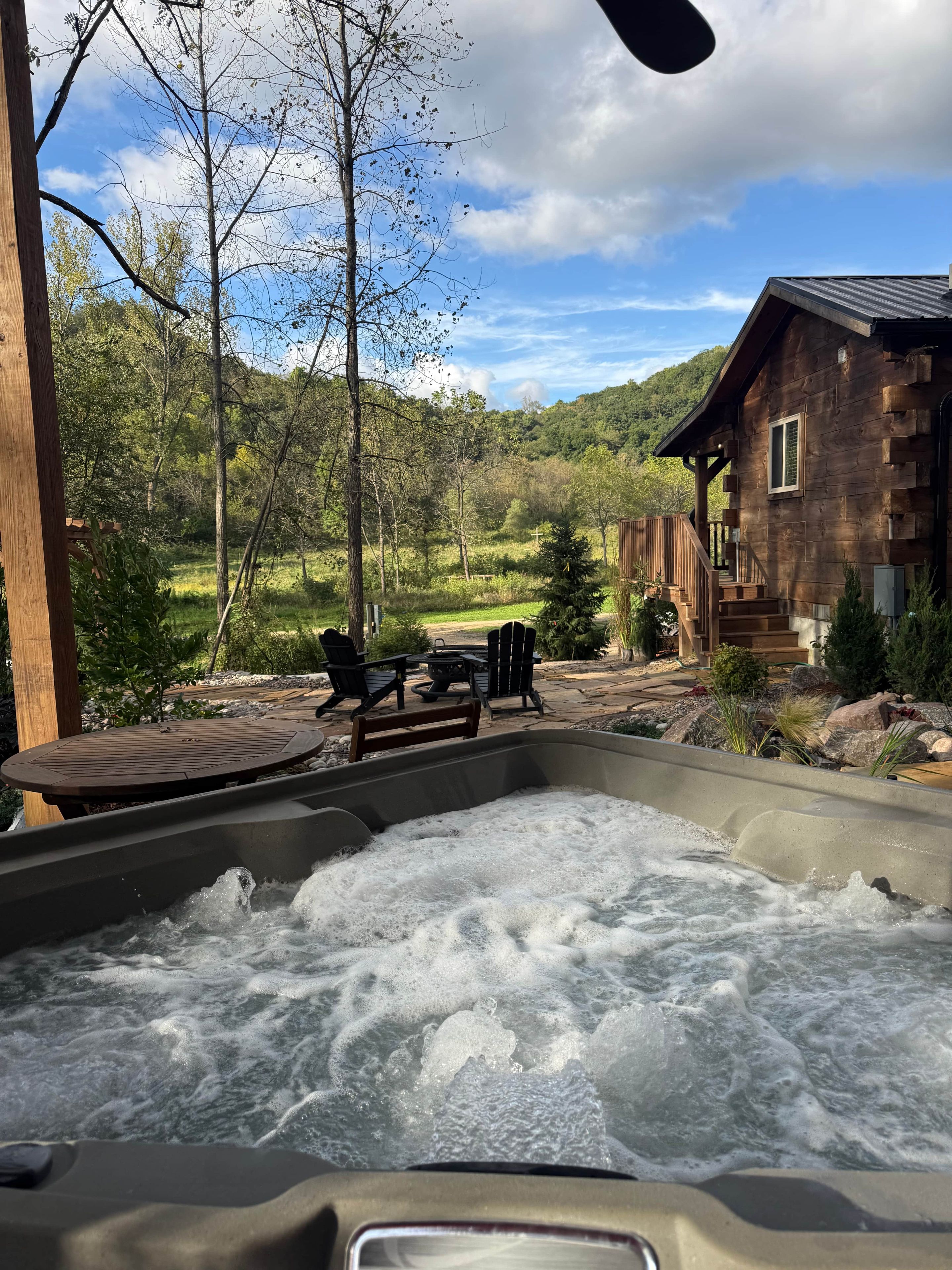 A hot tub with a wooden deck and outdoor seating, surrounded by a natural landscape and a wooden cabin.