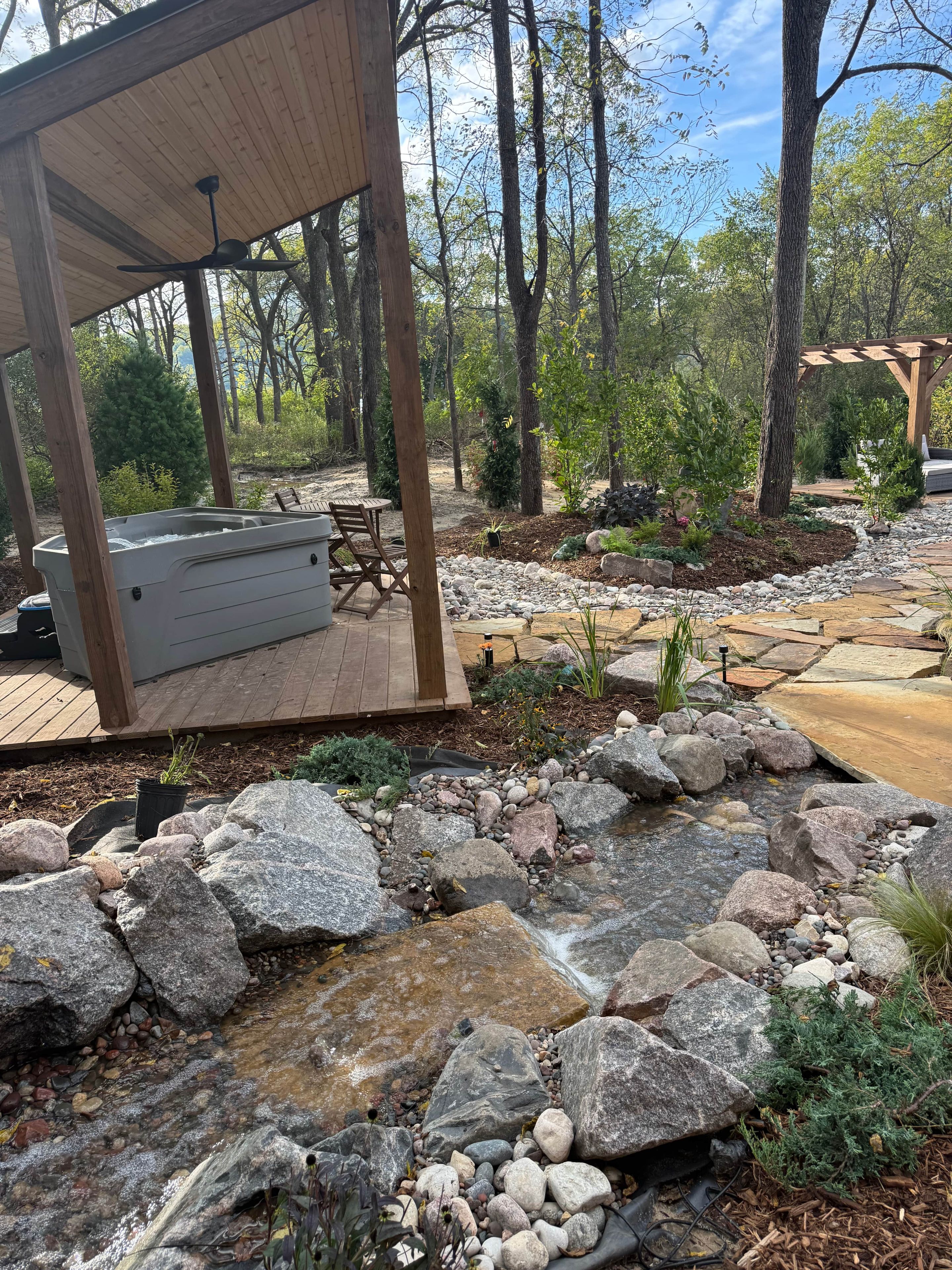 A covered outdoor seating area with a hot tub, surrounded by a natural landscape with trees and a small water feature.