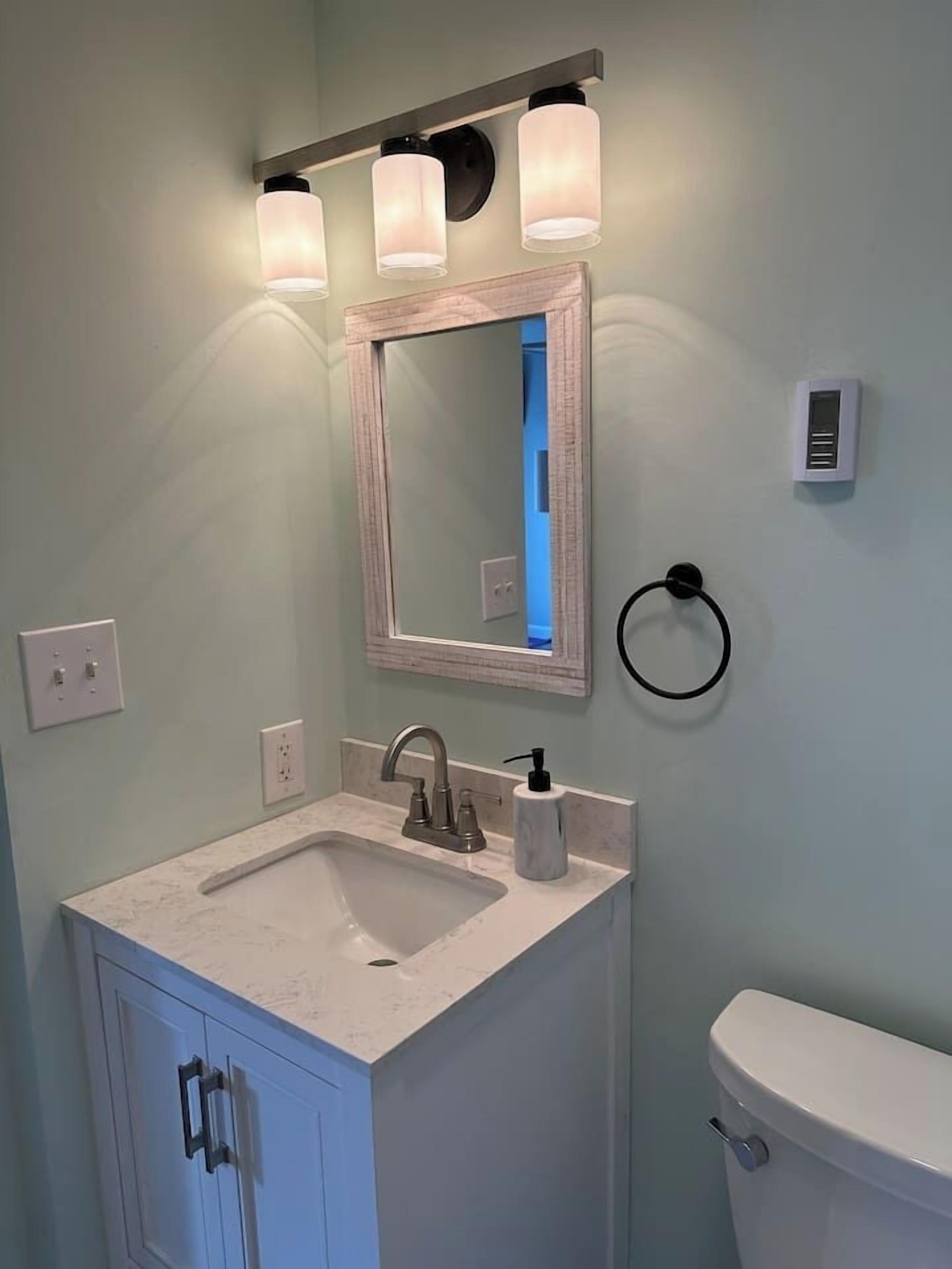 A bathroom with a white sink, a mirror, and a toilet. There are three wall-mounted lights above the mirror.