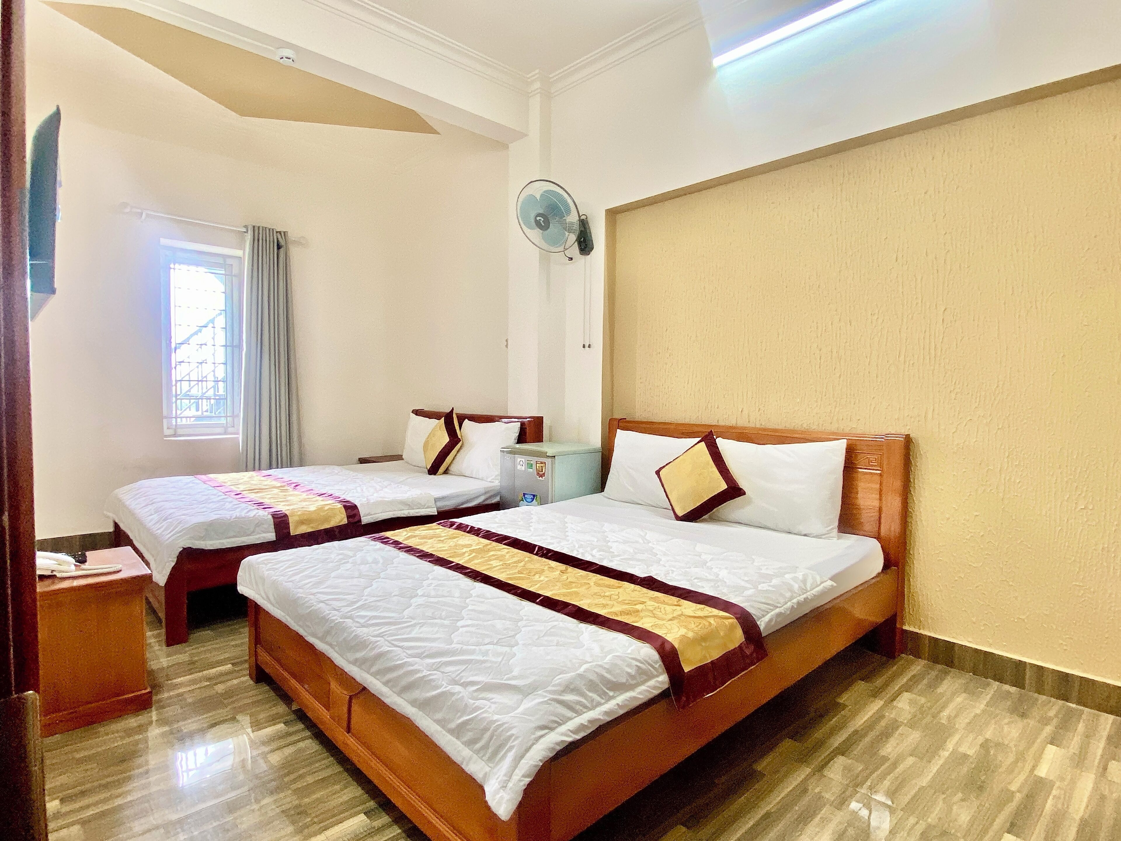 A hotel room with two beds, a bedside table, a nightstand, a window with curtains, and a wall-mounted fan.
