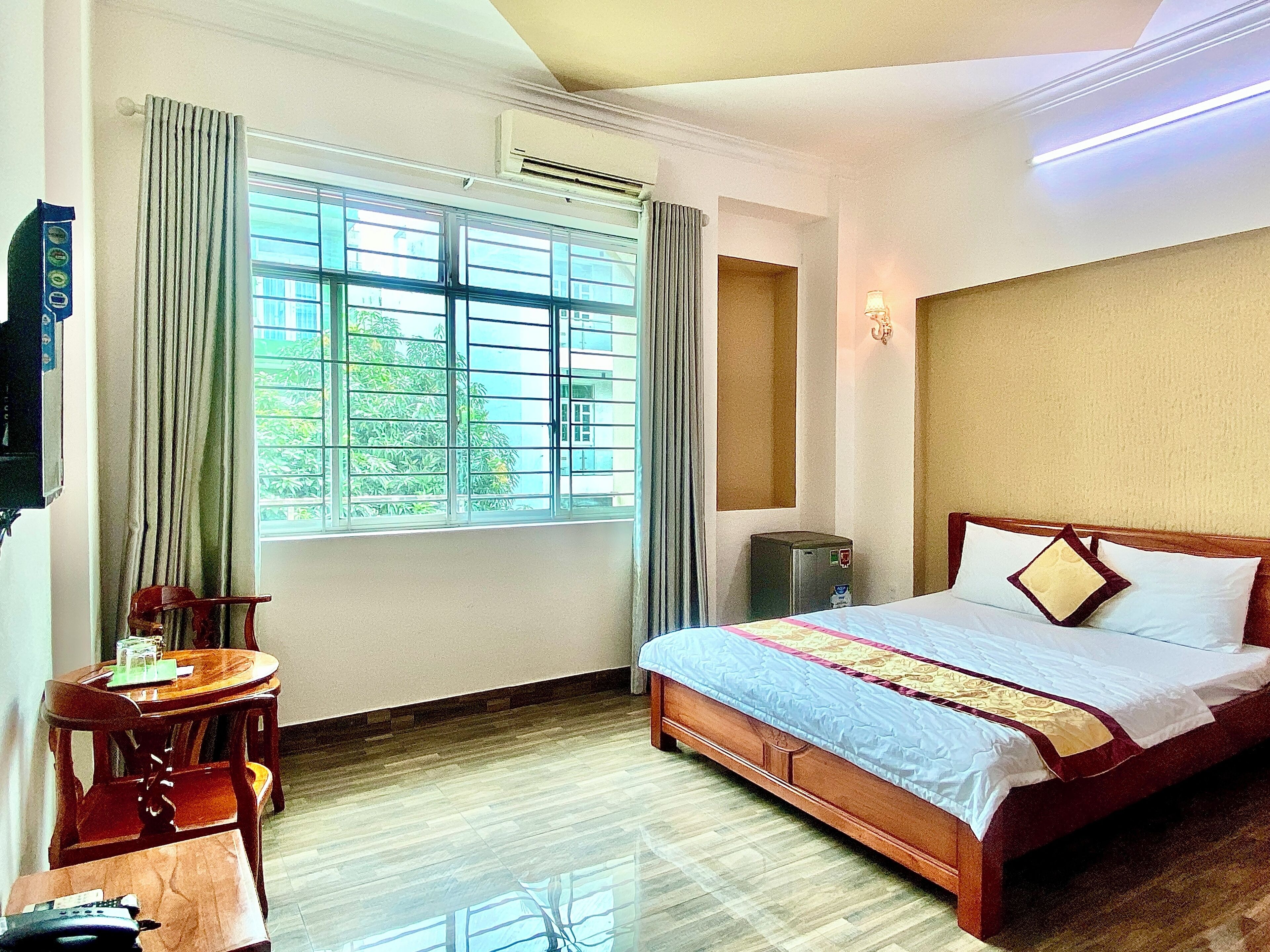 A hotel room with a wooden bed, a desk with a chair, a window with curtains, and an air conditioning unit.