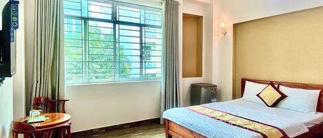 A hotel room with a wooden bed, a desk with a chair, a window with curtains, and an air conditioning unit.