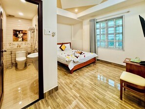 Ein Hotelzimmer mit einem hölzernen Bett, einem Badezimmer mit Toilette und Dusche, einem Schreibtisch mit Stuhl und einem Fenster mit Vorhängen.