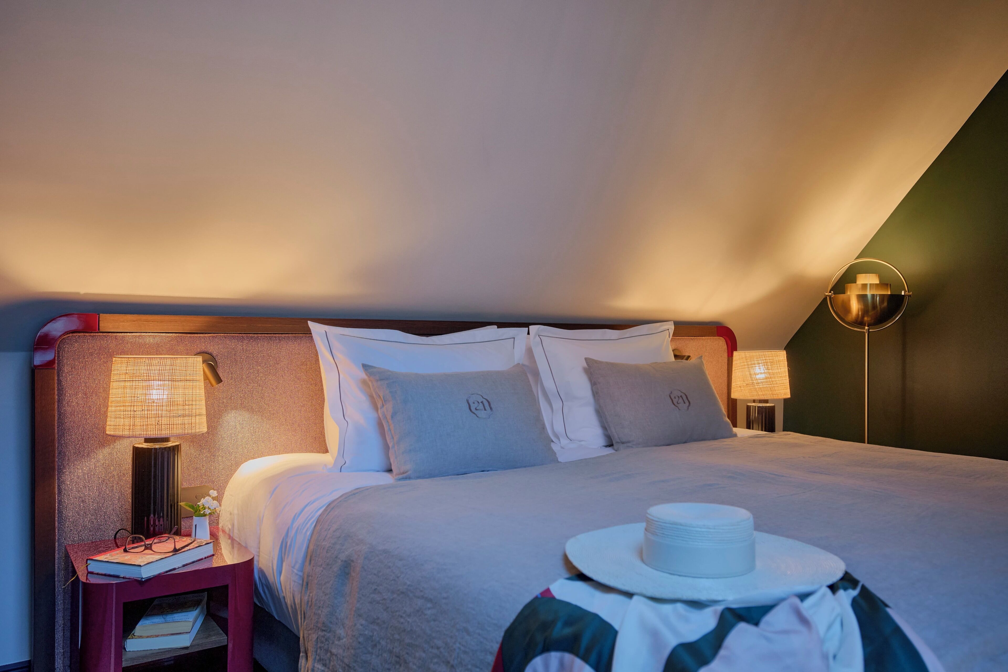 Un lit bien fait, avec un oreiller portant le chiffre « 21 », une lampe de chevet et une petite table sur laquelle se trouvent un chapeau et des livres.