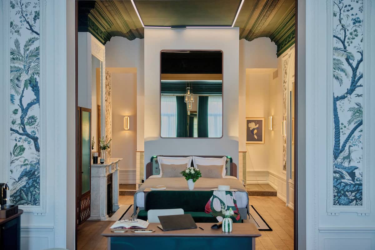 A modern hotel room with a large bed, a desk with a laptop, a vase with flowers, and a fireplace.