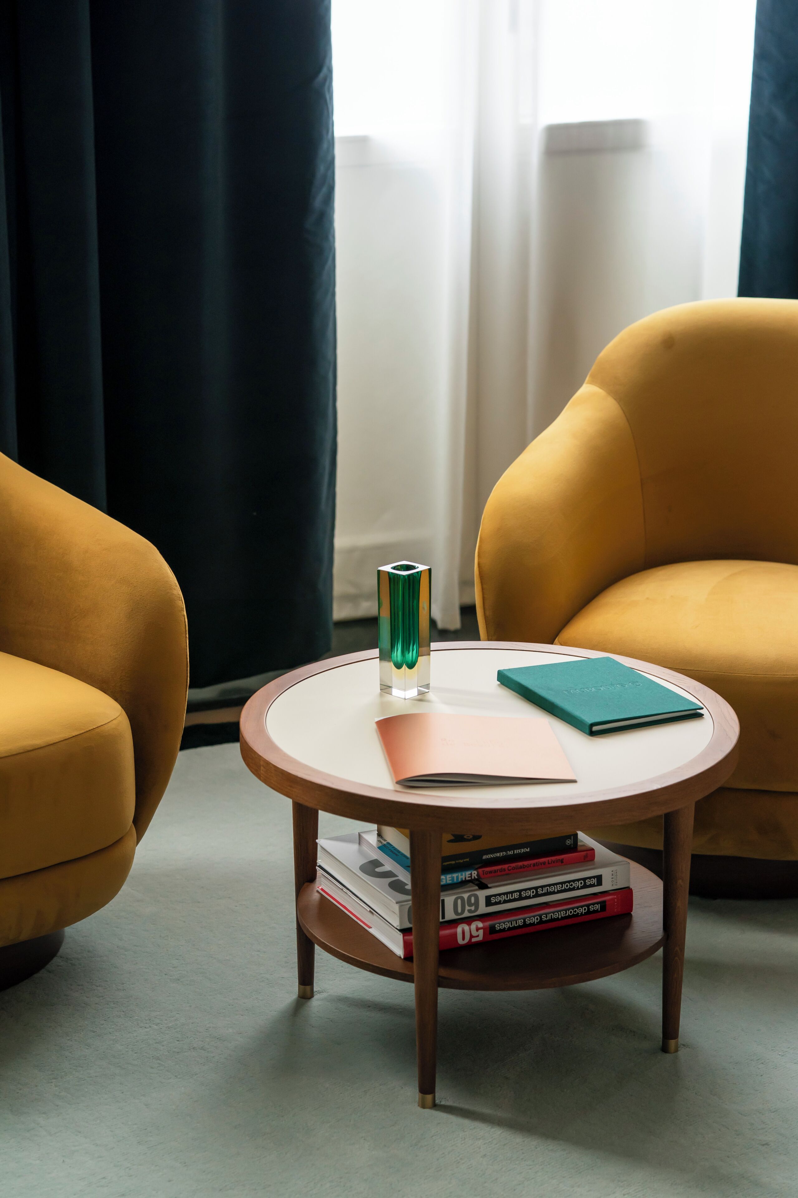 Een moderne woonkamer met een ronde salontafel, twee gele fauteuils, een boekenkast en een raam met gordijnen.