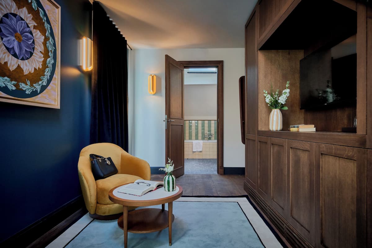A hotel room with a yellow armchair, a small round table with a book and a vase of flowers, a large floral artwork on the wall, a wooden cabinet, and a television on a wooden stand.