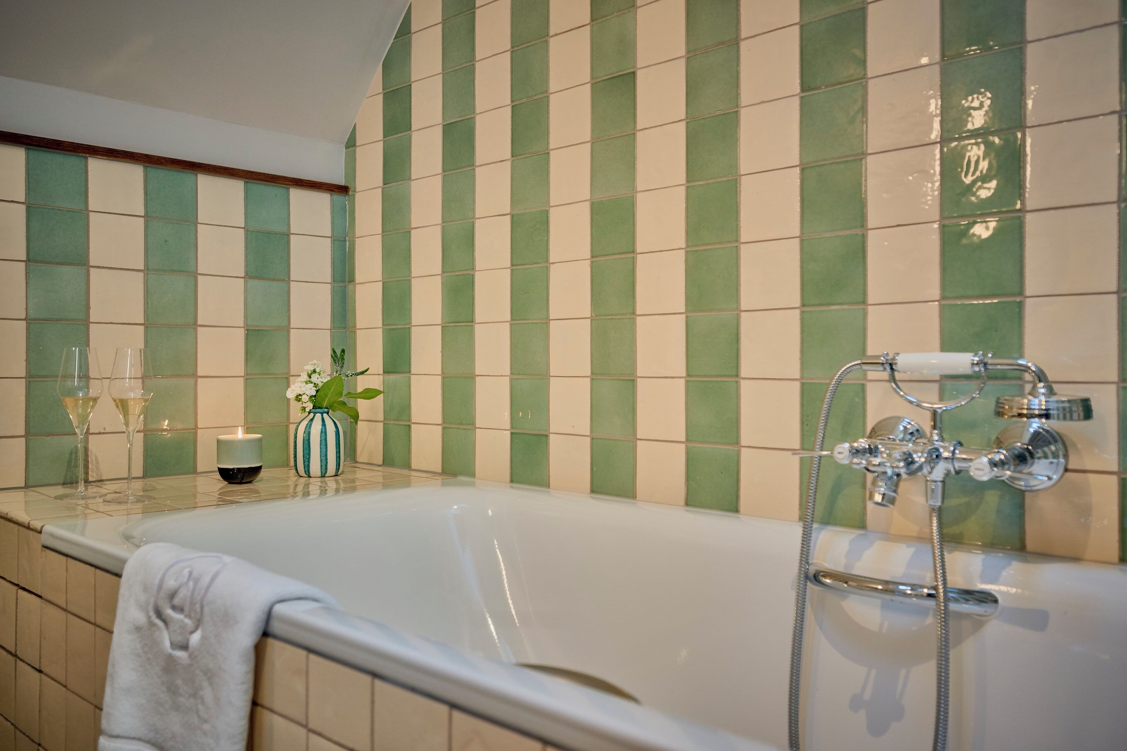 Een badkamer met een bad, groenbeige tegels, een glazen douchecabine en een vaas met bloemen op het aanrecht.