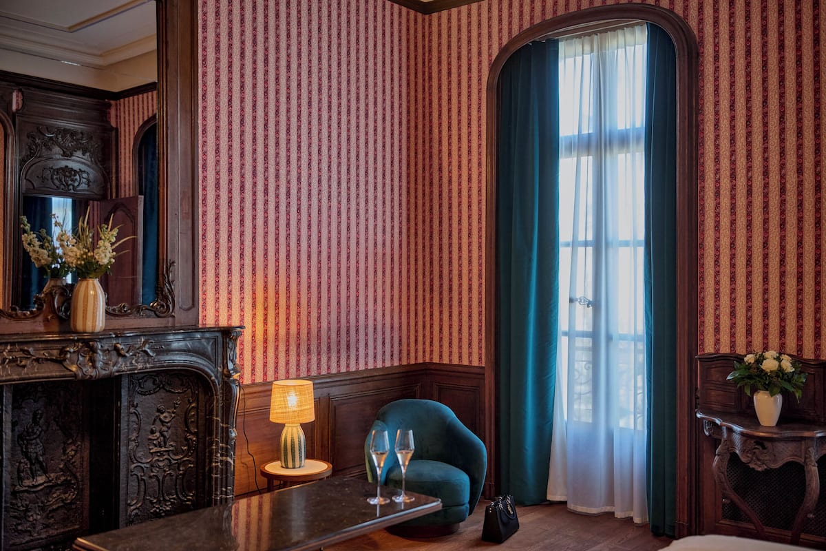 A room with striped wallpaper, a wooden fireplace, a table with a lamp, a blue armchair, and a window with curtains.