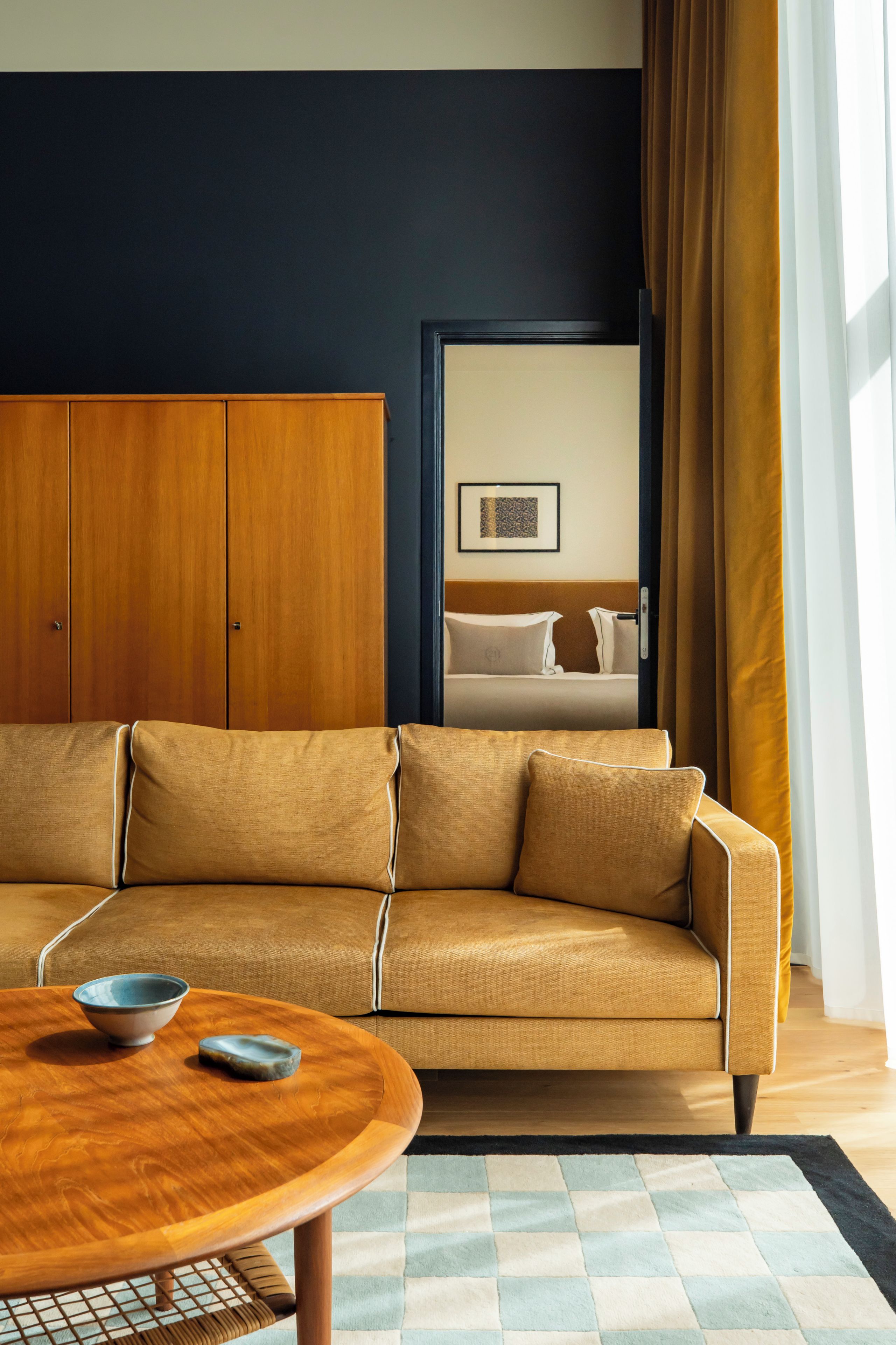 A modern living room with a sofa, a wooden coffee table, and a view of a bedroom with a bed and curtains.