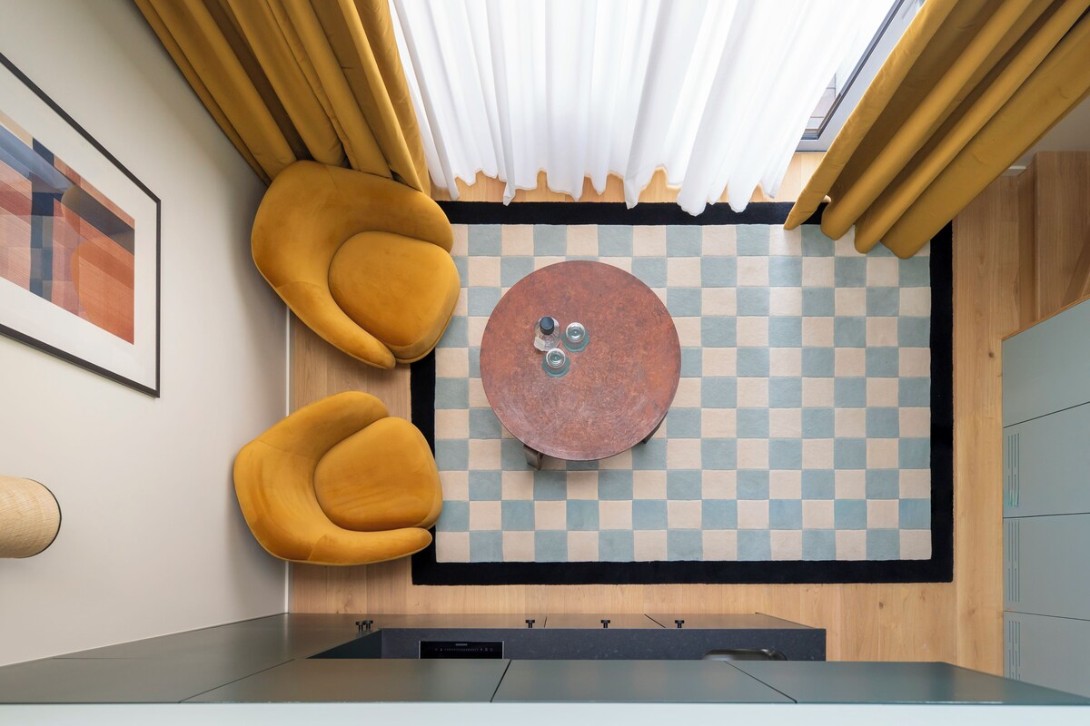 A modern room with a checkered floor, a round table with a copper top, and two mustard-colored armchairs.