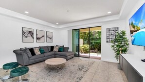 Uma sala de estar moderna com um sofá modular cinzento, uma mesa de centro redonda e uma poltrona verde. Há três quadros abstratos na parede, uma janela grande com vista para o verde e uma televisão fixada na parede.