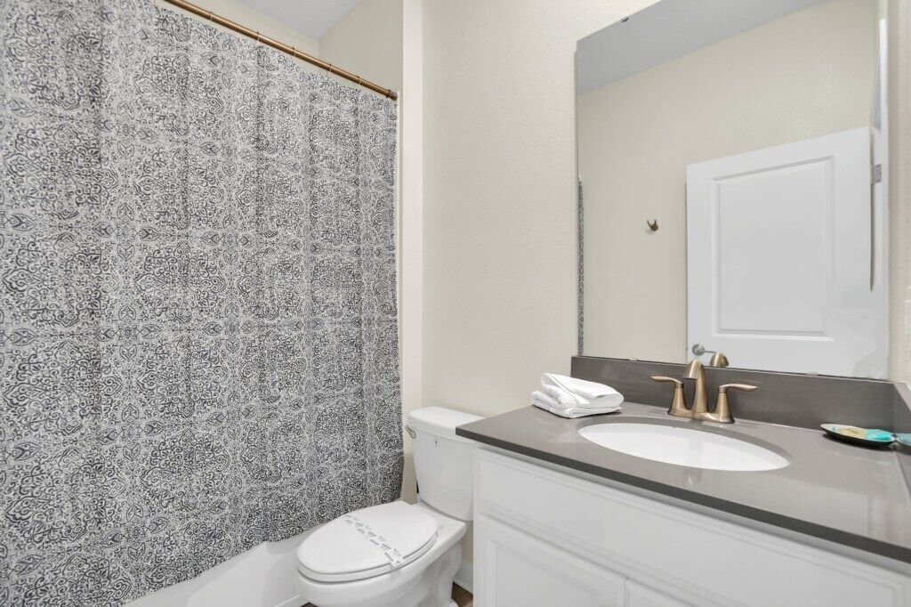 A bathroom with a patterned shower curtain, a white toilet, a sink with a gold faucet, and a mirror above the sink.