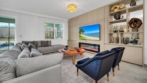 A modern living room with a large TV, a fireplace, and a view of the beach. - Storey Lake 9 BR Villa With Games Room (Kissimmee)