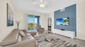 Ein modernes Wohnzimmer mit einem beigefarbenen Ecksofa, einem Glastisch und einem Flachbildfernseher, der an einer blauen Wand angebracht ist.