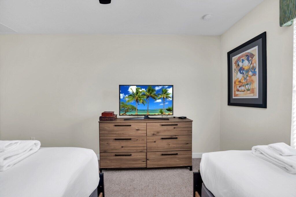A hotel room with two beds, a wooden dresser, a television displaying a tropical beach scene, and a framed cartoon character on the wall.