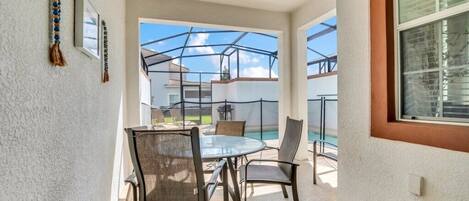 Un patio coperto con un tavolo e sedie in vetro, una piscina sullo sfondo e vista sulle case vicine.