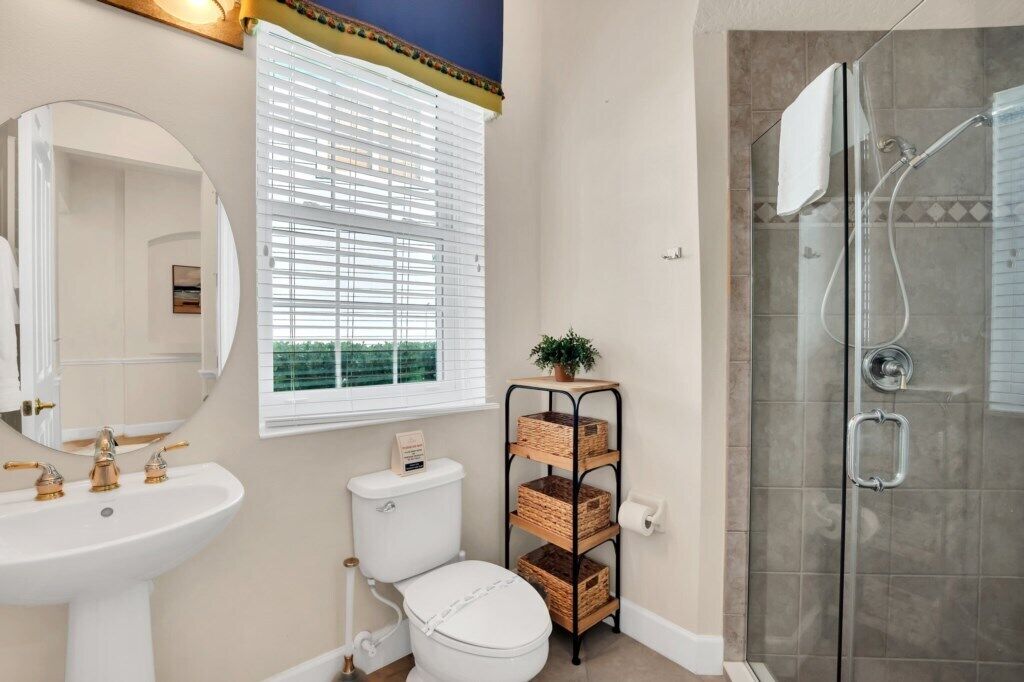 A bathroom with a white sink, a toilet, a mirror, a window with blinds, a shower enclosure, and a wicker shelf with baskets.