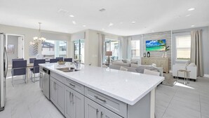 A modern kitchen with a large island, dining area with chairs, and a living room with a sofa and TV.