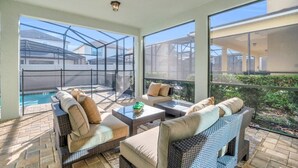 A sunlit patio with wicker furniture, a pool, and large windows offering views of the surrounding area.