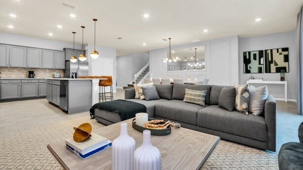 A modern living room with a grey sofa, coffee table, and decorative items. A kitchen with grey cabinets and a dining area with a white table and chairs are visible.