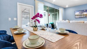 A dining area with a wooden table set for four, a vase of purple flowers, and a view of a living room with a sofa and a painting on the wall. - Ultimate 5 Bedroom Windsor Island Resort Pool  home (Davenport)