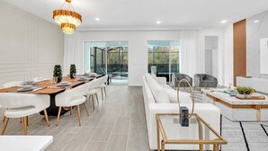 Een moderne eetruimte met een lange tafel, witte stoelen en een kroonluchter. Er is een zithoek met een bank en fauteuils.