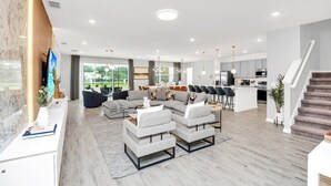 A modern living room with a sectional sofa, a coffee table, and a TV. There is a kitchen area with a dining table and chairs.