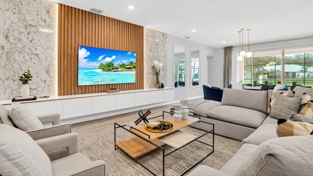 A modern living room with a large flat-screen TV, a wooden accent wall, and a view of a tropical beach through large windows.