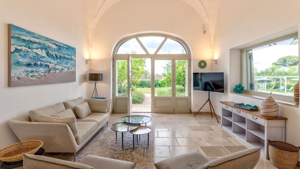 A spacious living room with a high arched doorway leading to a garden, a comfortable sofa, a glass coffee table, and a flat-screen TV mounted on the wall.