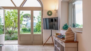 A room with a large arched window, a flat-screen TV on a tripod stand, a small cactus, and a woven basket on a wooden shelf.