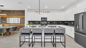 Cocina moderna con armarios blancos, una isla grande con taburetes de bar, un refrigerador de acero inoxidable e iluminación colgante.