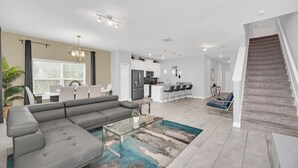 Una sala de estar moderna con un sofá modular, una mesa de centro de vidrio y un comedor con mesa y sillas. Se observa una cocina al fondo con una barra y zona de estar. Una escalera es visible a la derecha.