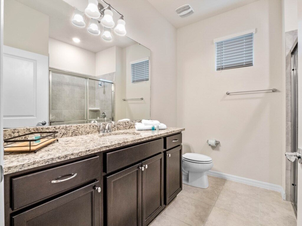 A bathroom with a granite countertop, dark cabinets, a white toilet, and a glass shower enclosure.