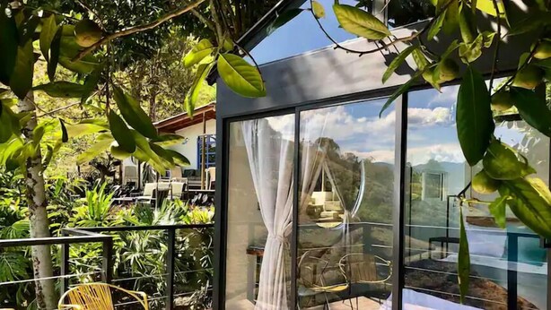 A modern outdoor seating area with glass walls, a view of a mountainous landscape, and lush greenery. - La Comarca Guatape (El Peñol)