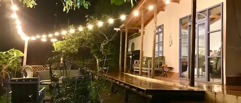Un espacio de estar al aire libre techado, con luces colgantes, un deck de madera y un edificio con grandes ventanales.