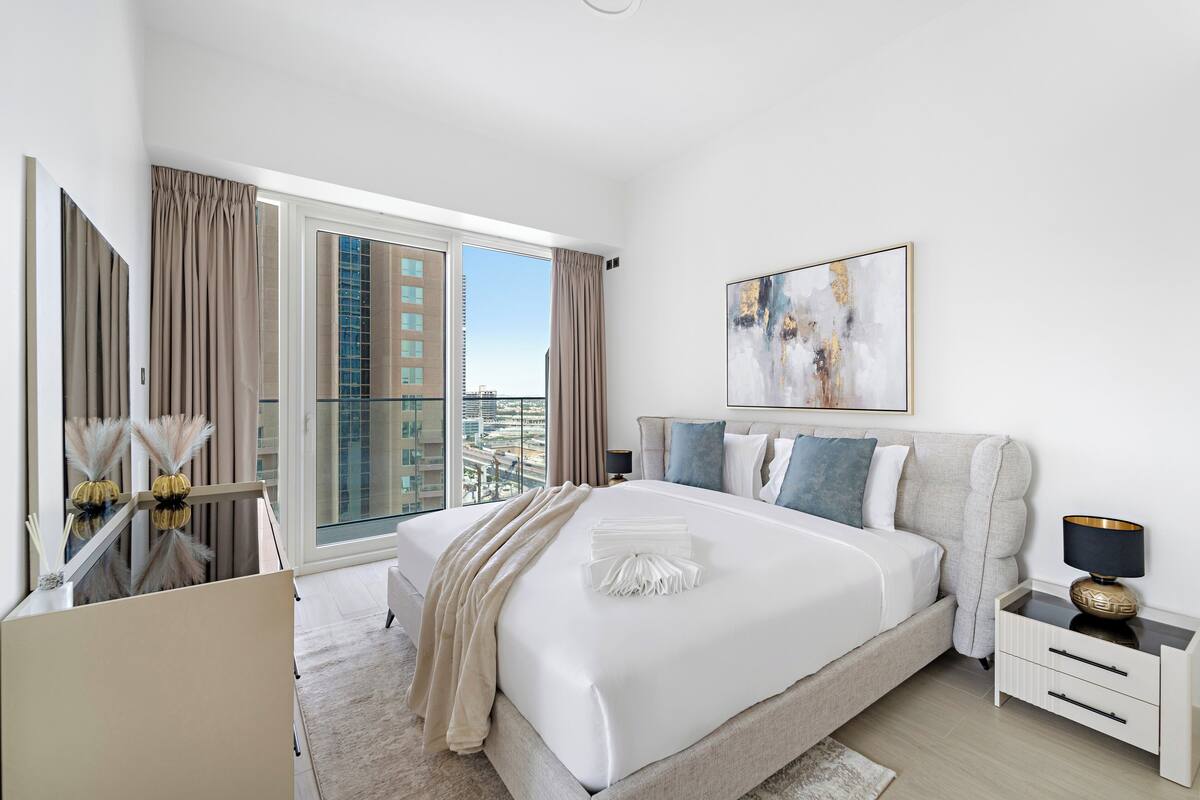 A modern hotel room with a large bed, a nightstand, a desk, and a view of the cityscape through floor-to-ceiling windows.