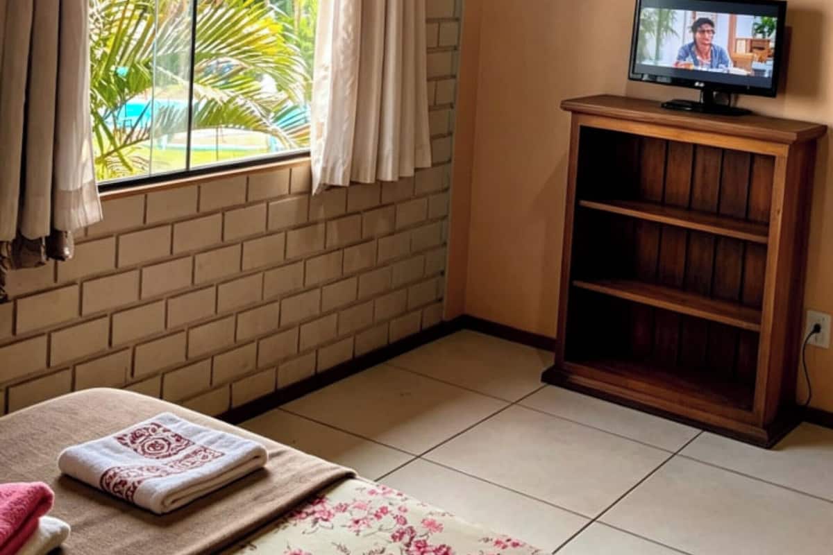 A room with a brick wall, wooden ceiling, a TV on a wooden stand, a bed with a floral bedspread, and a window with curtains.