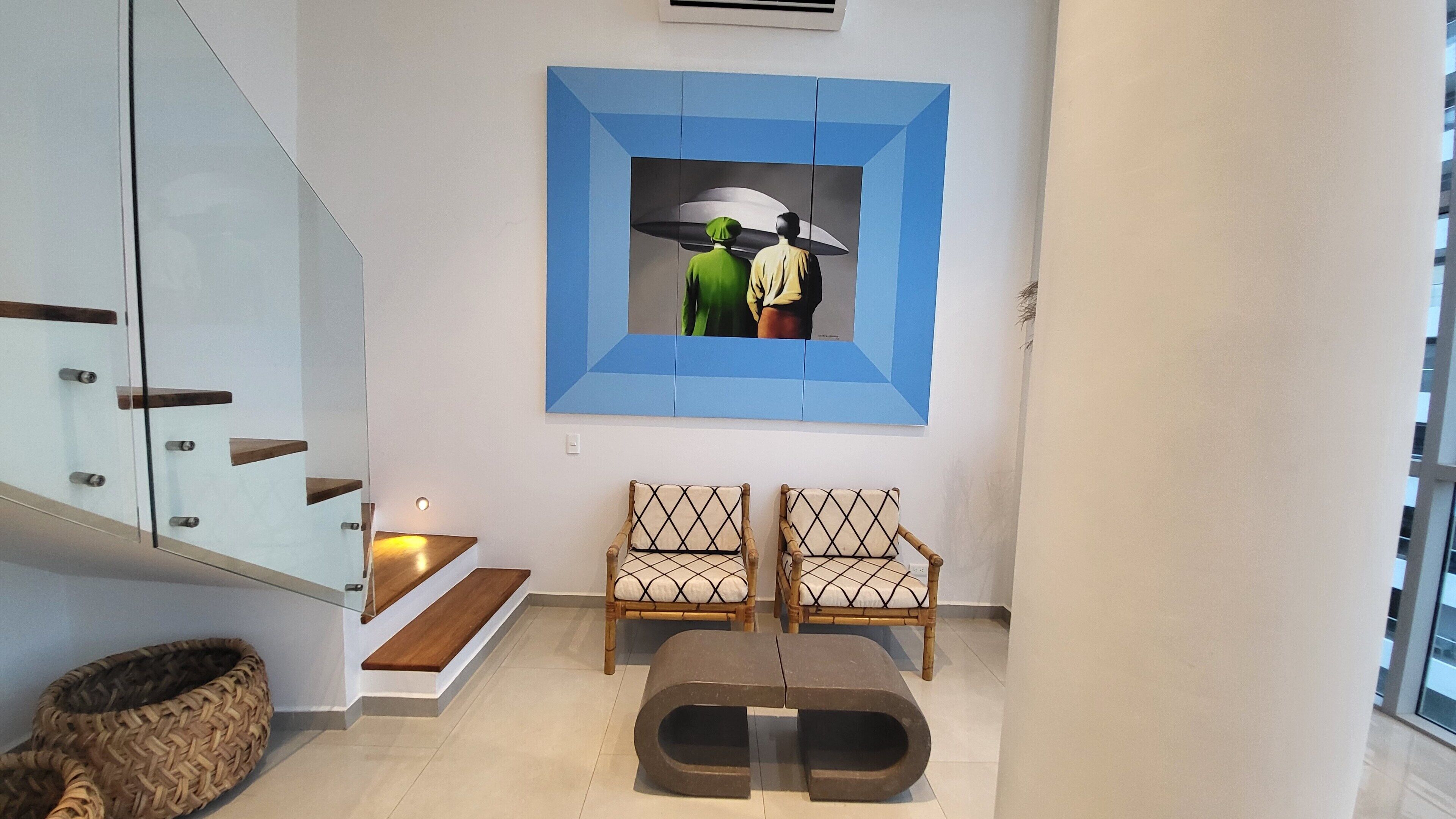 Ein modernes Wohnzimmer mit Treppe, zwei Sesseln, einem Couchtisch und einem an der Wand hängenden Gemälde.