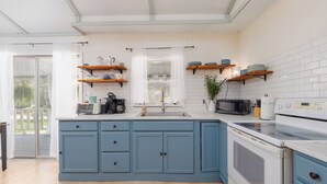 A modern kitchen with blue cabinets, white countertops, and a large island. There are wooden shelves above the counters, and a window with a view of greenery outside. - Satilla Woods Cabin & Hot Tub (Hortense)