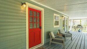 A porch with a red door, two chairs, a small table, and a welcome mat. - Satilla Woods Cabin & Hot Tub (Hortense)