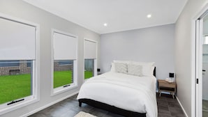 Quarto com cama, mesa de cabeceira, luminária e janelas amplas.