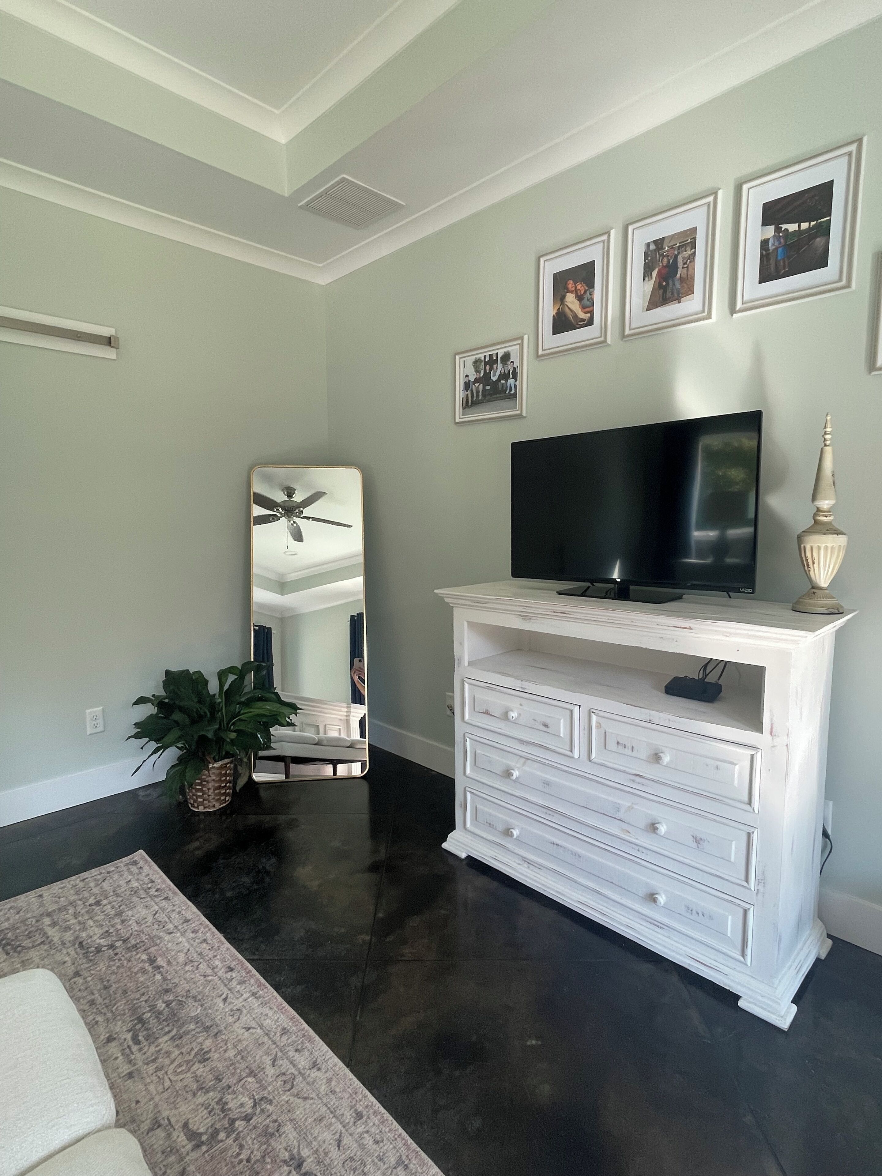 A living room with a dark floor, a white dresser with drawers, a flat-screen TV, a large mirror, and framed pictures on the wall.
