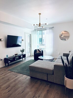 A modern living room with a flat-screen TV, a round mirror, a ceiling fan, and a large window with curtains. - Modern and Updated 2-bed/2-bath Condo in Silicon Valley! (Los Angeles)