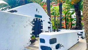 Un edificio blanco con una reja negra, rodeado de palmeras y un edificio rojo al fondo.