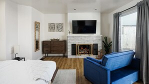 A modern bedroom with a large bed, a blue sofa, a fireplace, and a TV mounted above it. - Ocean Beach Spanish Gem | Walk to beach pier shops (San Diego)