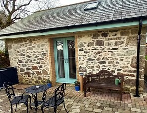 Un edificio de piedra con una puerta color turquesa, un pequeño patio con mesa y sillas, y un banco de madera.